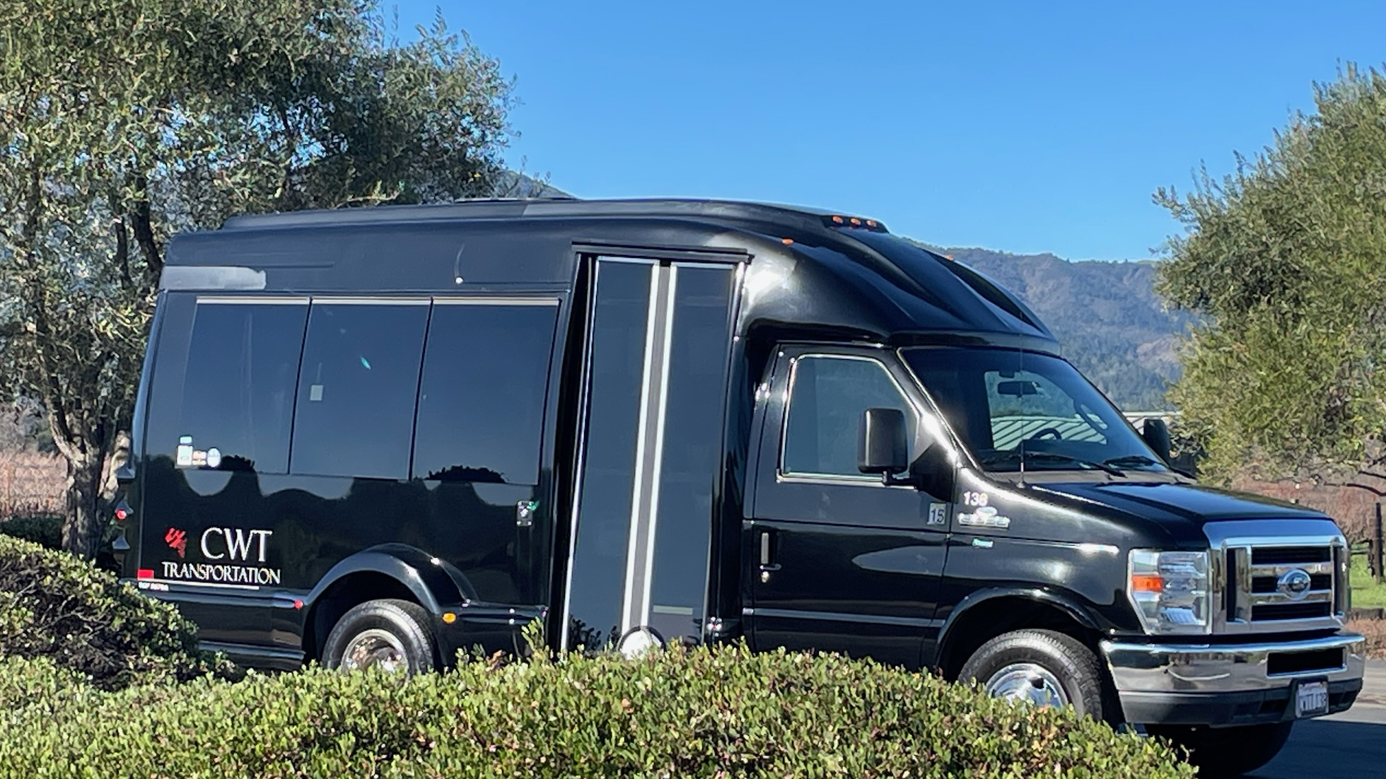 California Wine Tours black CWT shuttle bus parked by vineyard with scenic mountain backdrop for group wine tours